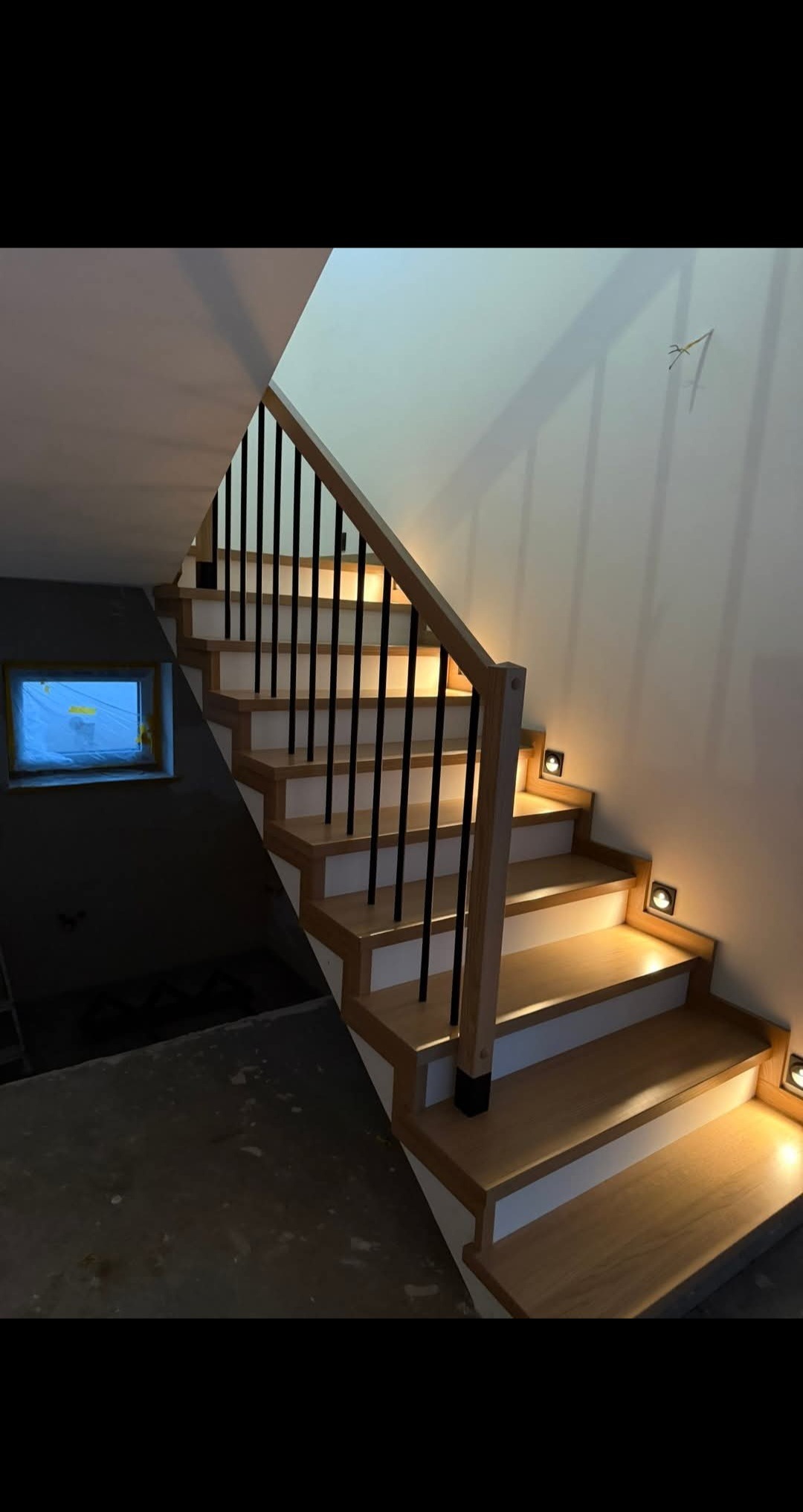 Jasne schody z drewnianymi stopniami, czarną balustradą i subtelnym oświetleniem punktowym. Widoczne okno w surowym stanie. Ujęcie z dołu, podkreślające detale.