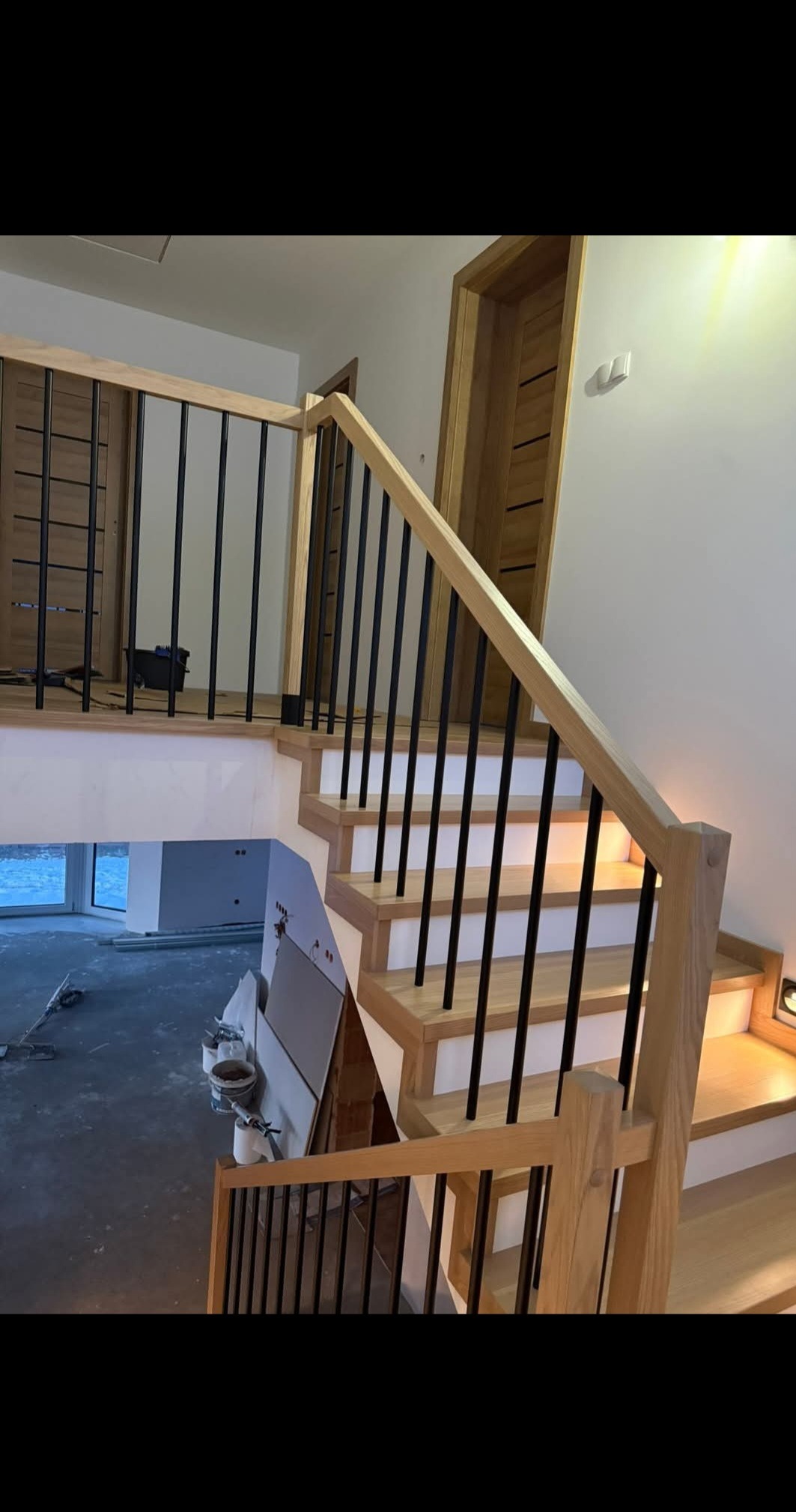 Drewniane schody z czarną balustradą w nowoczesnym wnętrzu. Widoczne detale wykończenia i oświetlenie schodów. W tle drzwi z drewnianymi akcentami.