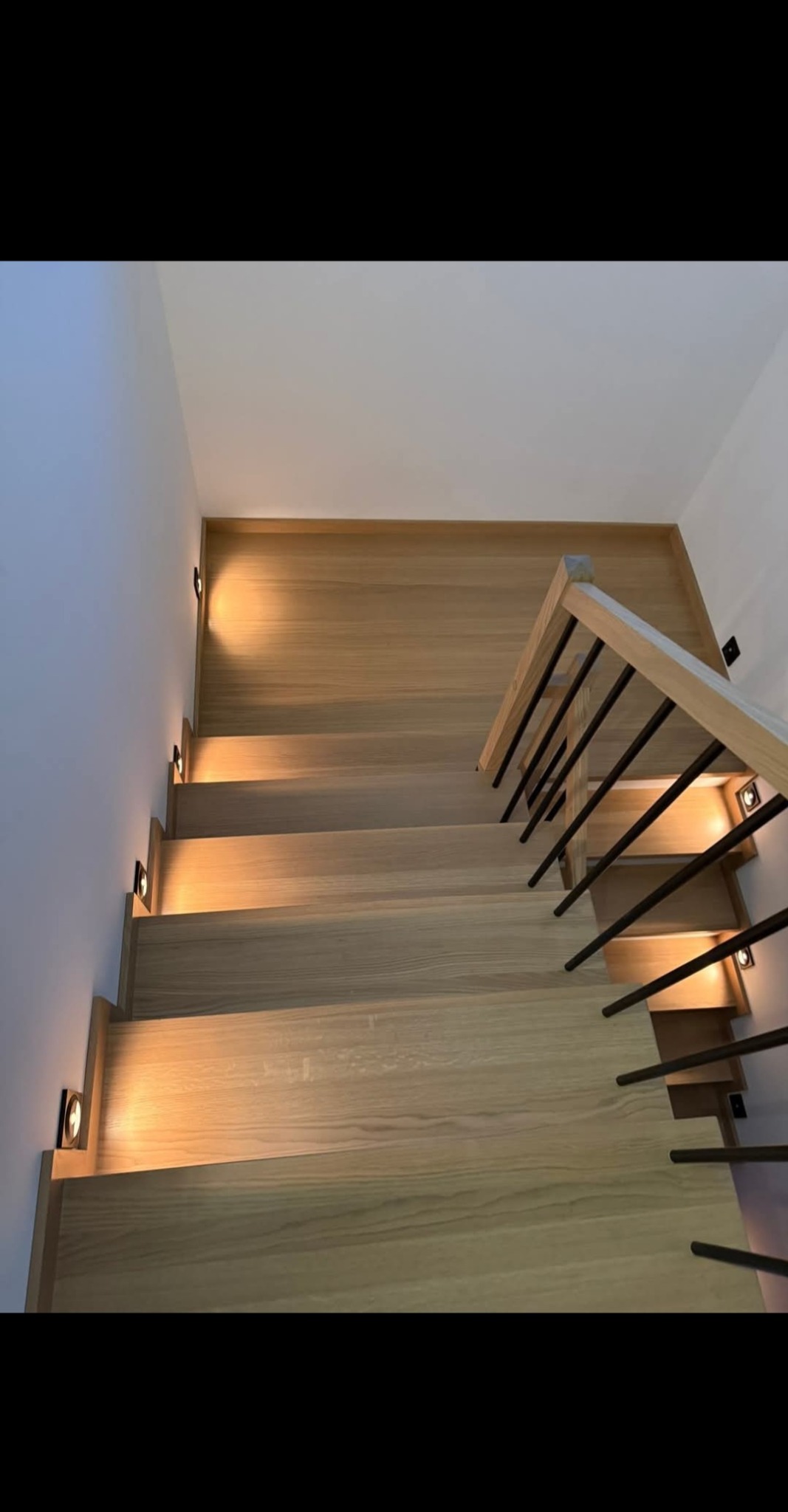 Jasne, drewniane schody z minimalistyczną balustradą i subtelnym oświetleniem punktowym wzdłuż ściany, widziane z góry.