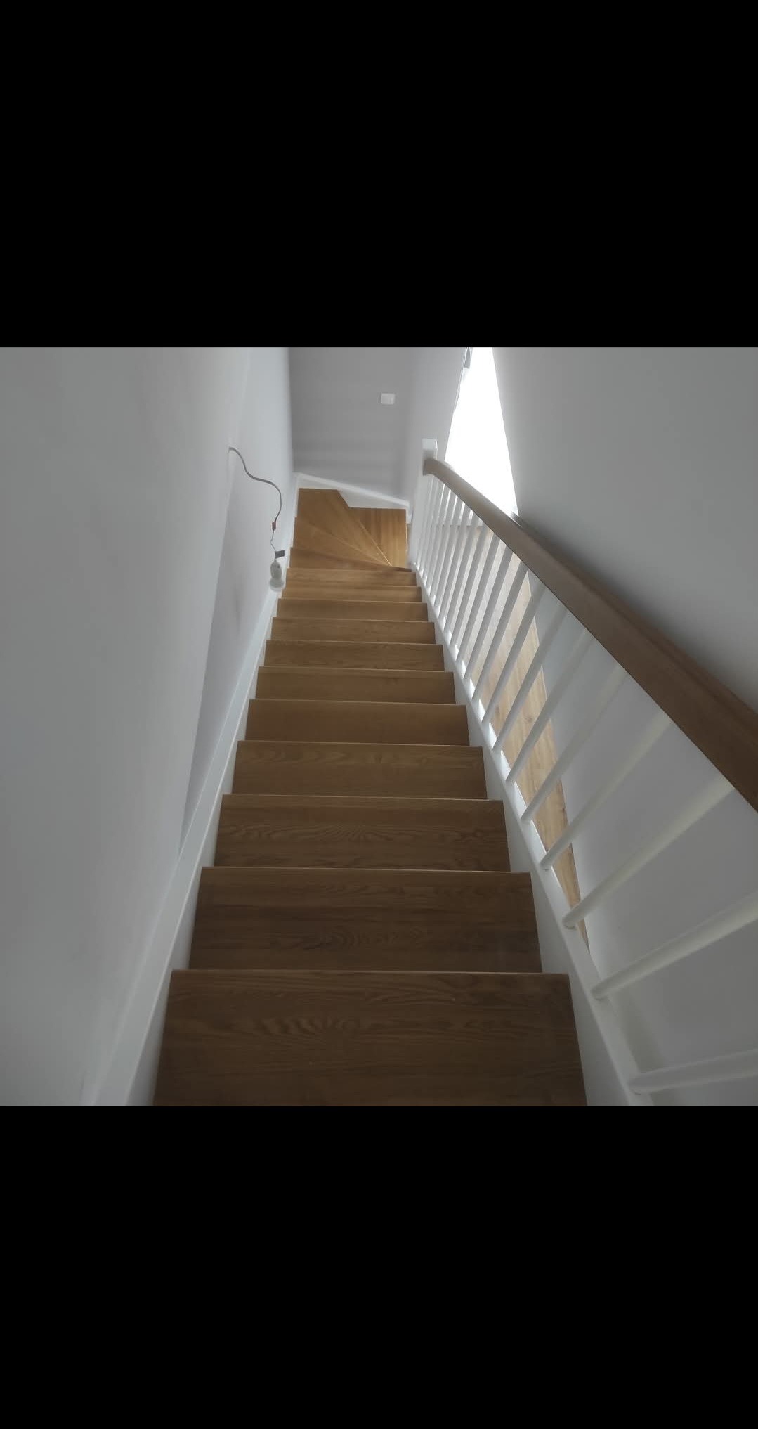 Jasne, drewniane schody z białą balustradą, widziane z dołu. Minimalistyczny design, naturalne światło wpadające z góry. Widoczny fragment instalacji elektrycznej.
