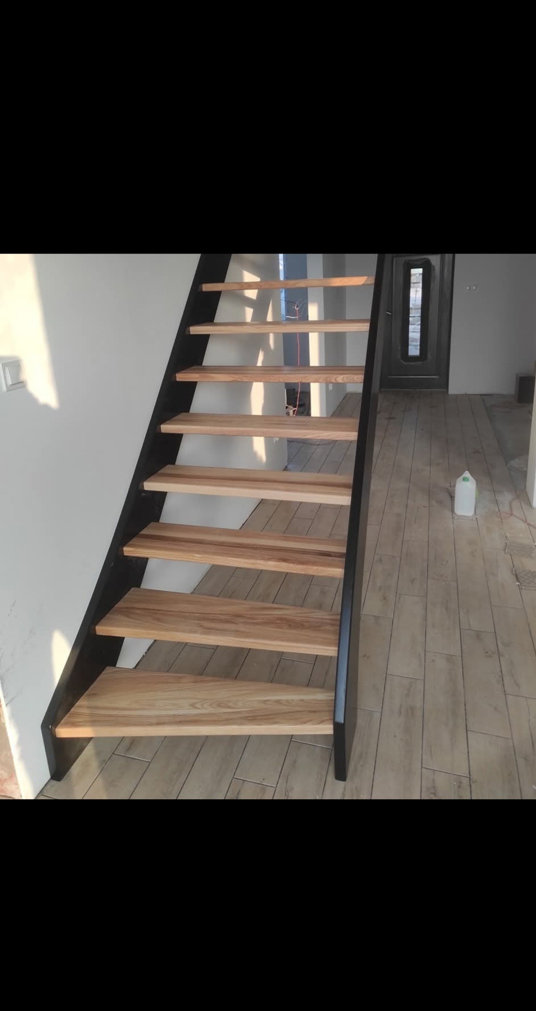 Nowoczesne schody z drewnianymi stopniami i czarną metalową konstrukcją, widok z dołu, jasne wnętrze w trakcie wykończenia, butelka wody na podłodze.
