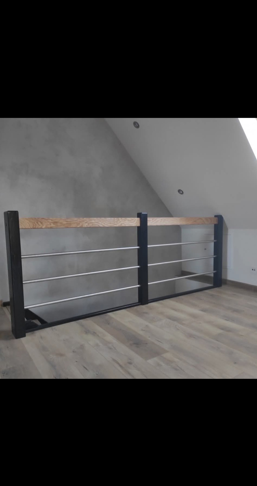 Nowoczesna balustrada z czarnymi słupkami, drewnianą górną belką i metalowymi prętami, zamontowana w jasnym wnętrzu poddasza z drewnianą podłogą.