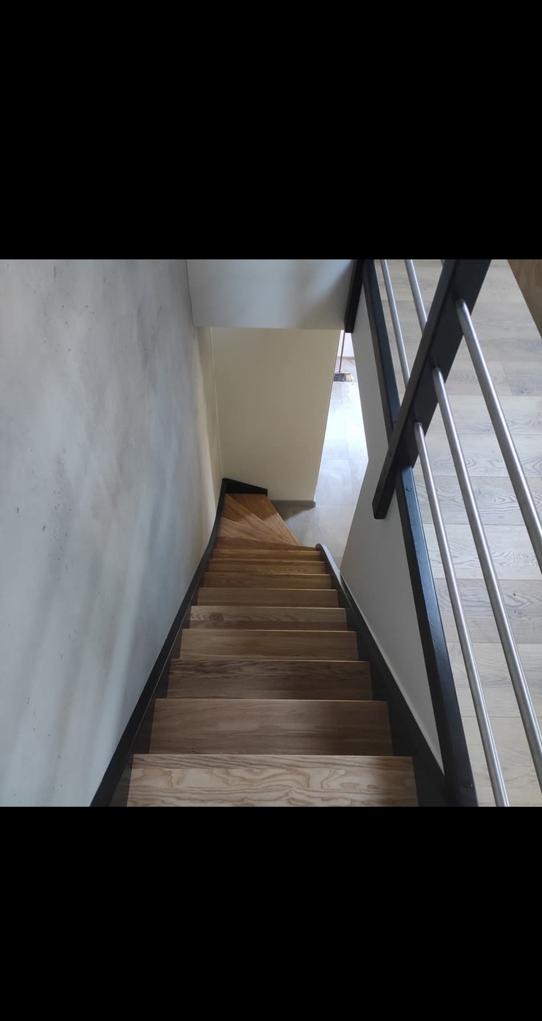 Drewniane schody z metalową balustradą, widok z góry. Stopnie z jasnego drewna, ciemne obramowanie. Minimalistyczny design, szara ściana i jasna podłoga na górze.