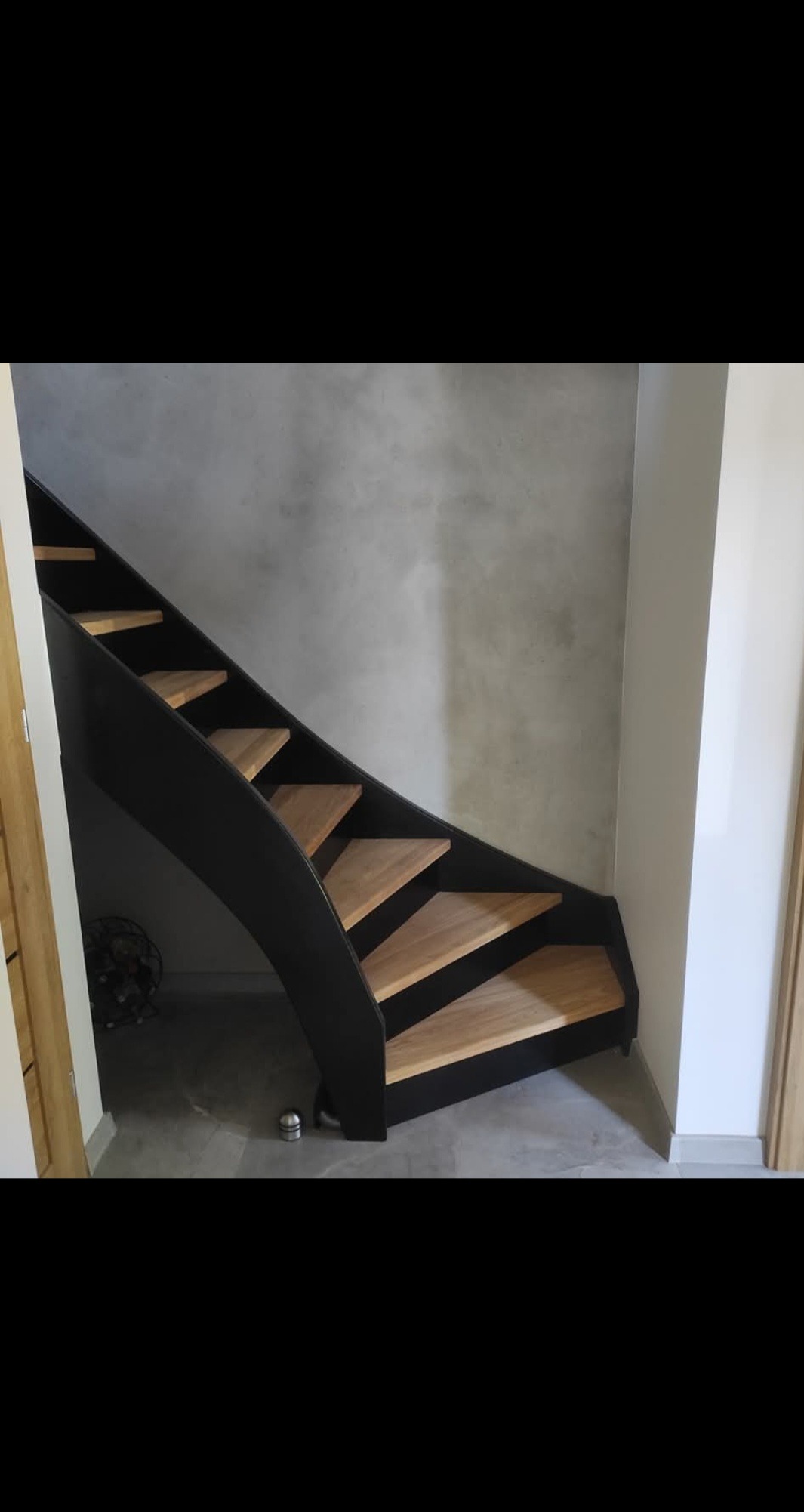 Nowoczesne schody z drewnianymi stopniami i czarną, giętą konstrukcją, widoczne z dołu, na tle szarej ściany. Minimalistyczny design i precyzyjne wykonanie.