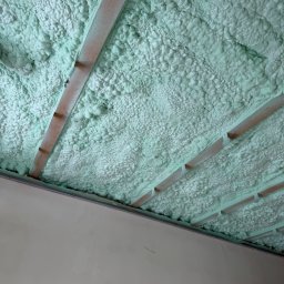 BIRTECH JAKUB BIR - Poddasze ocieplone pianką PUR w kolorze jasnozielonym, widoczne drewniane elementy konstrukcji dachu. Aplikacja pianki wykonana precyzyjnie, wypełniając całą przestrzeń.