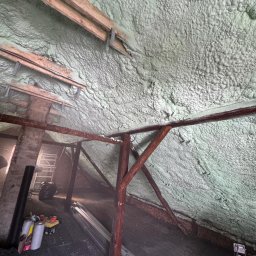Poddasze z drewnianą konstrukcją pokryte szarozieloną pianą PUR, widoczne narzędzia i materiały budowlane. Ujęcie z dołu, ukazujące strukturę izolacji.