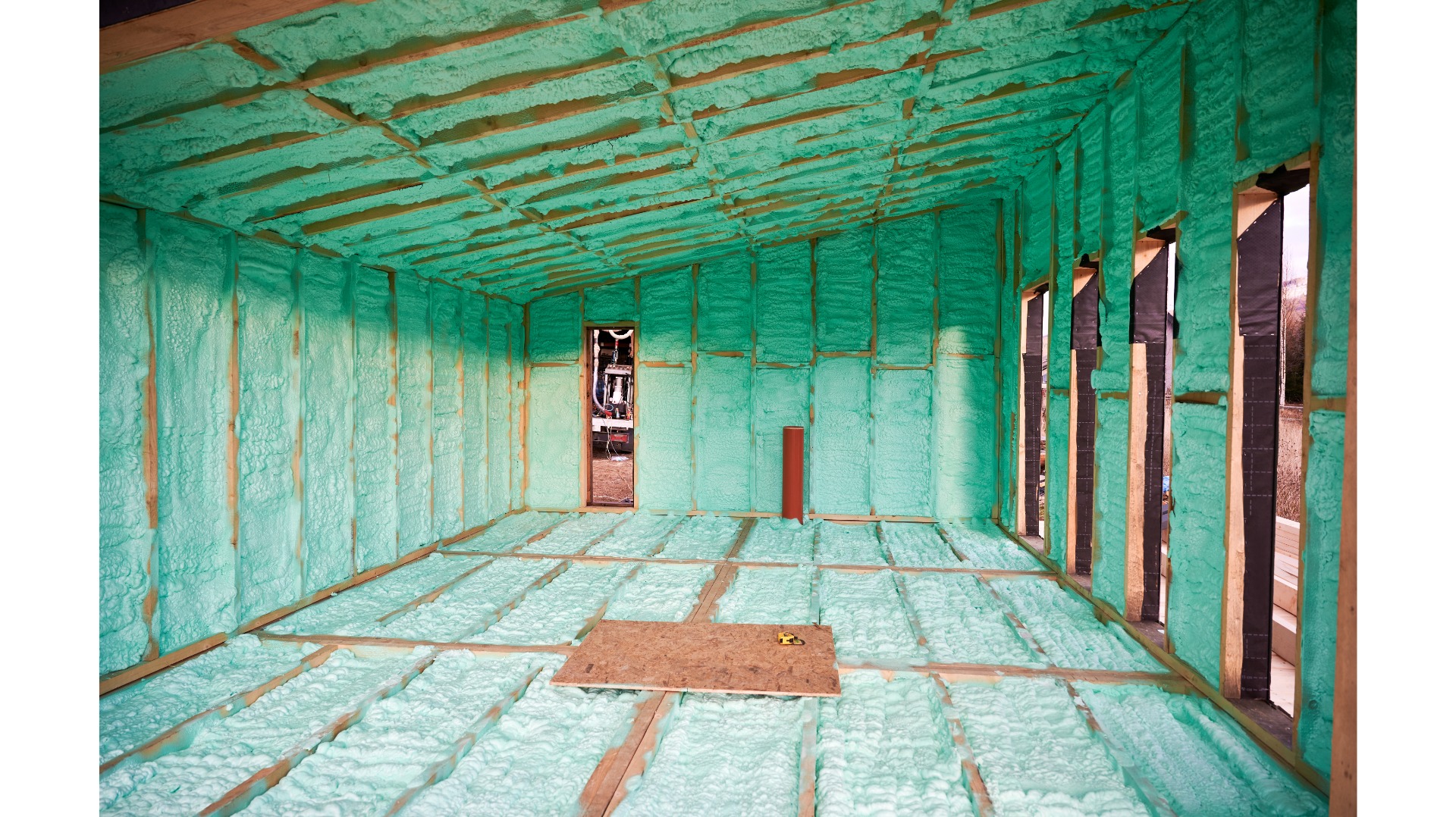 Poddasze domu w trakcie ocieplania pianką PUR, widoczne drewniane elementy konstrukcyjne i zielona pianka izolacyjna. Na podłodze płyta i miarka.