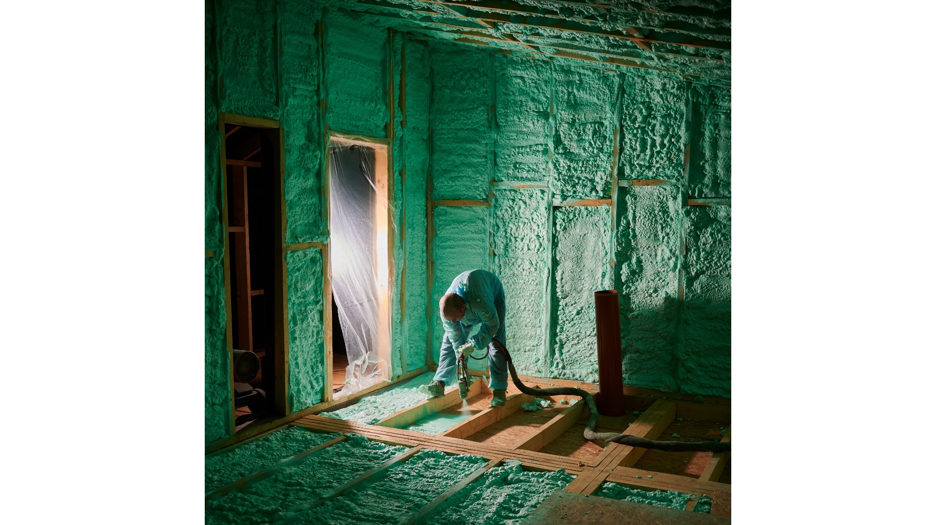 Pracownik w kombinezonie aplikuje piankę PUR na poddaszu, widoczne drewniane elementy konstrukcyjne i częściowo ocieplone ściany. Ochrona folią przy drzwiach.