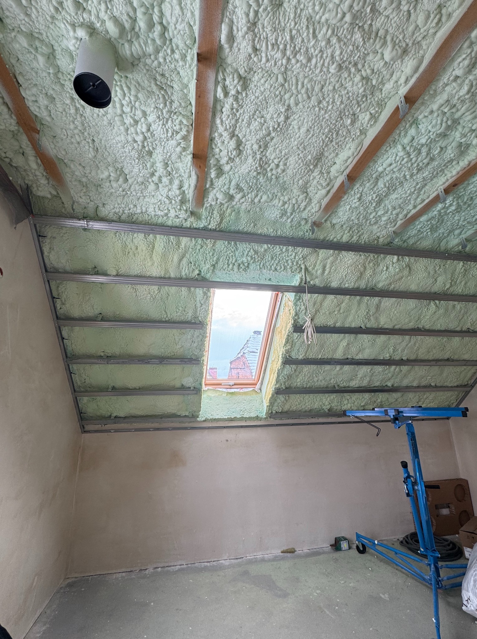 Poddasze po natrysku pianki PUR, widoczne drewniane belki, metalowe profile, okno dachowe i niebieski podnośnik do płyt. Świeżo wykonana izolacja.