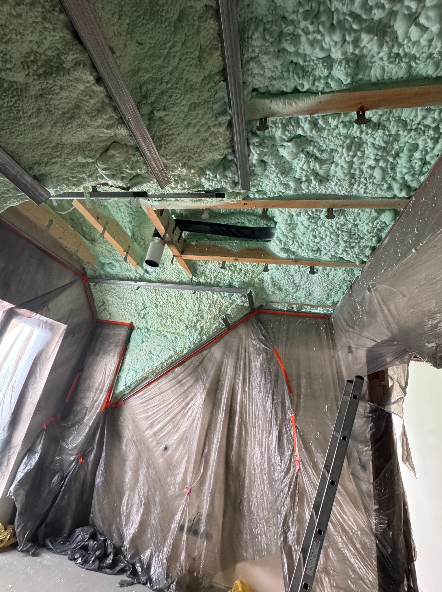 Poddasze z widoczną strukturą pianki izolacyjnej PUR, zabezpieczone folią ściany i sufit, drabina oparta o ścianę. Widoczne elementy konstrukcji drewnianej i metalowej.