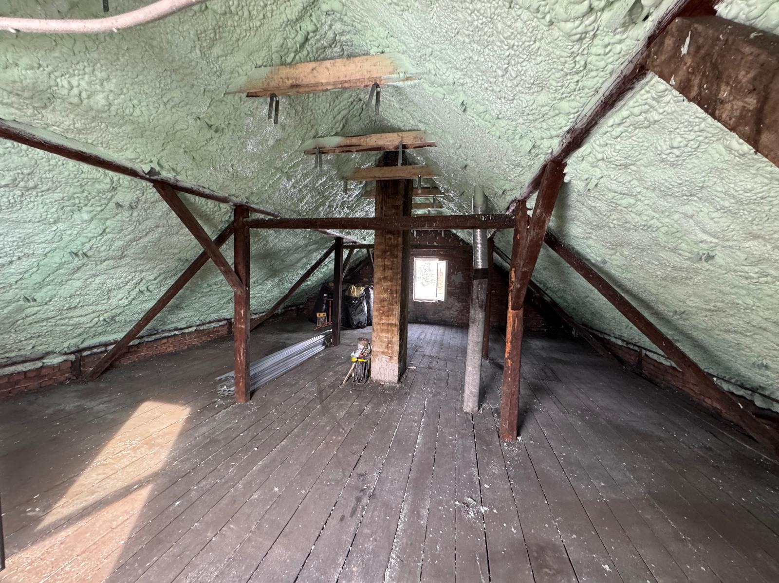 Poddasze z drewnianą konstrukcją i ociepleniem pianką PUR na skośnych ścianach. Widoczne narzędzia i materiały budowlane. Małe okno w tle.