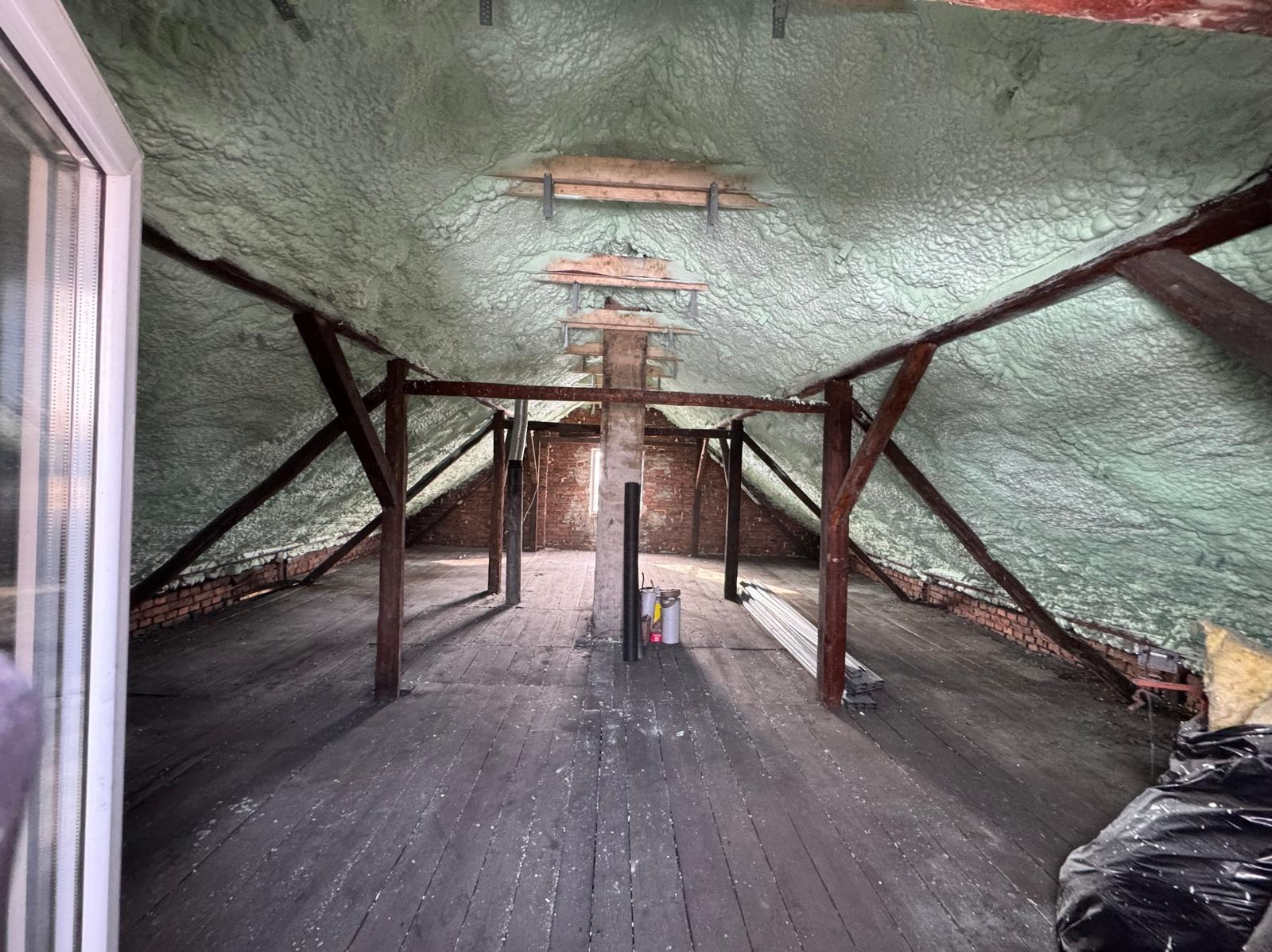 Poddasze z drewnianą konstrukcją i ceglanymi ścianami po aplikacji izolacji pianką PUR. Widoczne nierównomierne pokrycie pianką i elementy konstrukcyjne.