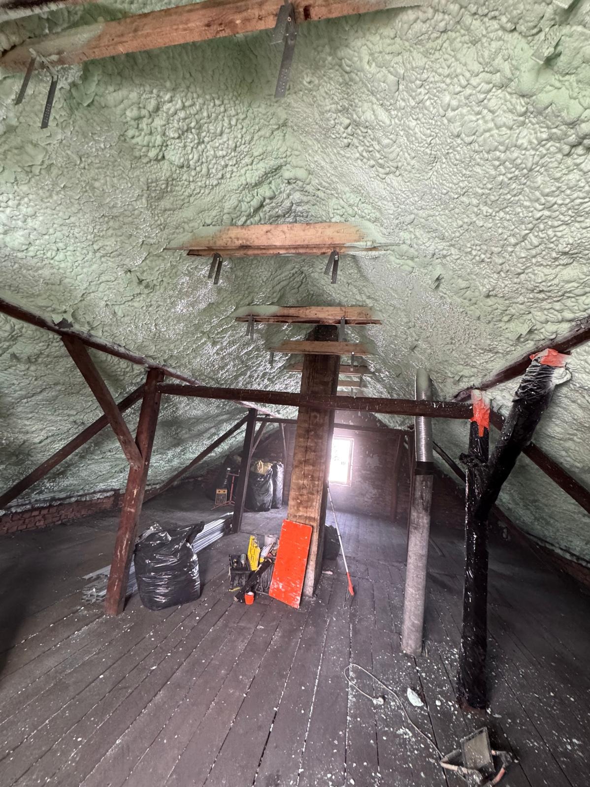 Poddasze po ociepleniu pianką PUR, widoczne drewniane elementy konstrukcyjne i warstwa izolacji. Na podłodze narzędzia i worki. Światło wpada przez okno.