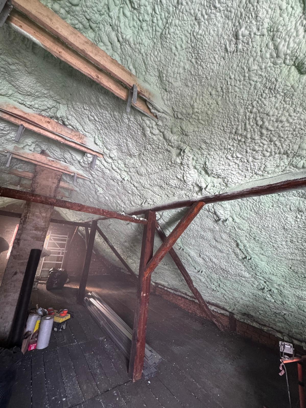 Poddasze z drewnianą konstrukcją pokryte szarozieloną pianą PUR, widoczne narzędzia i materiały budowlane. Ujęcie z dołu, ukazujące strukturę izolacji.