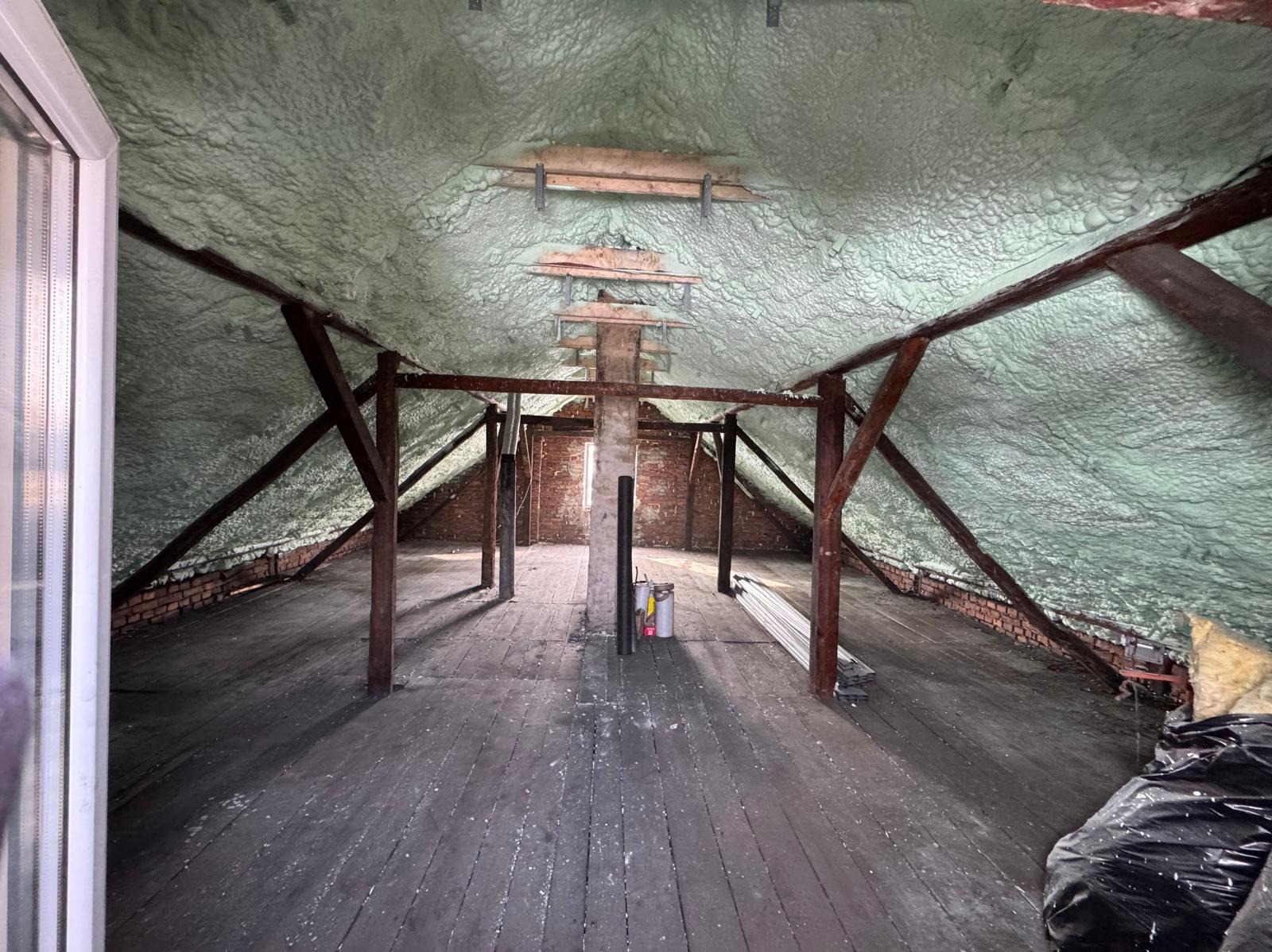 Poddasze z widoczną konstrukcją drewnianą i zaaplikowaną pianą izolacyjną na skośnych powierzchniach dachu. Widoczne ceglane ściany i drewniana podłoga.