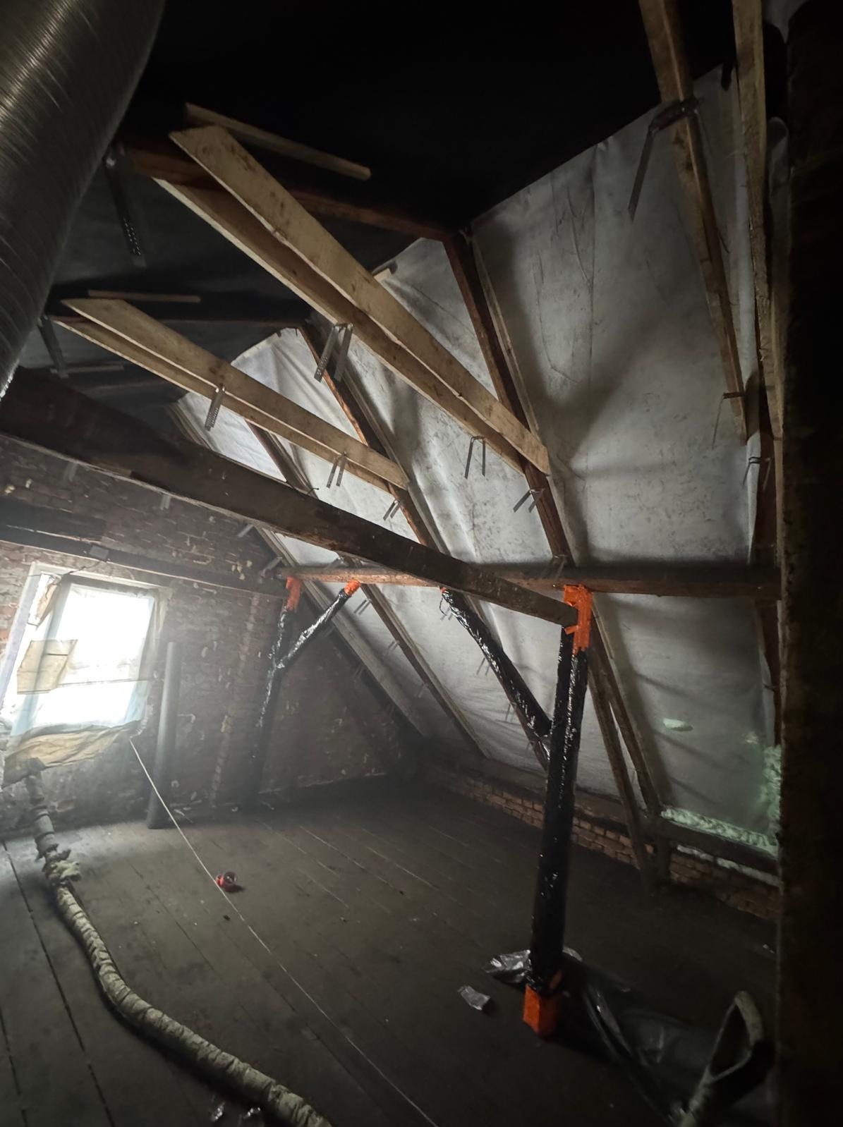 Poddasze w trakcie prac izolacyjnych: widoczne drewniane krokwie, folia paroizolacyjna i elementy montażowe. Częściowo ocieplone ściany szczytowe z cegły.