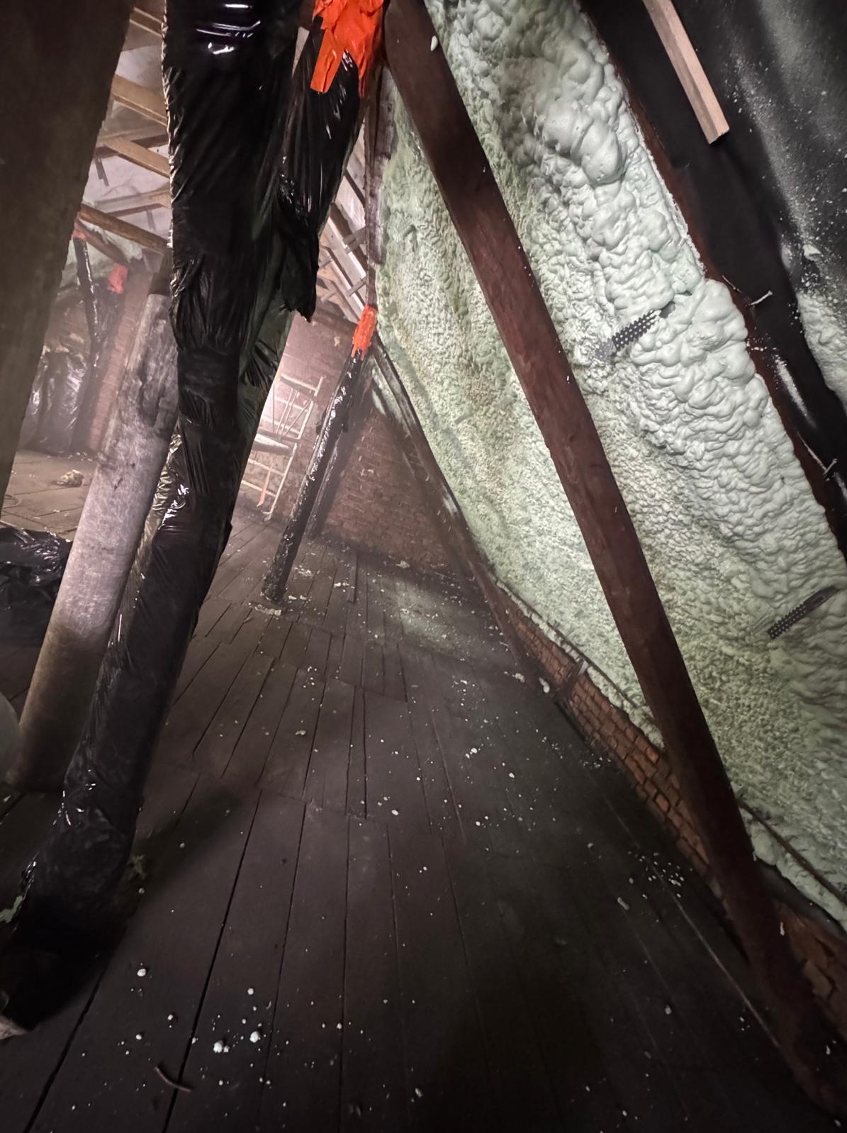 Poddasze z drewnianą konstrukcją, częściowo pokryte jasną pianą izolacyjną. Elementy konstrukcji zabezpieczone folią. Widoczne drewniane deski podłogowe.