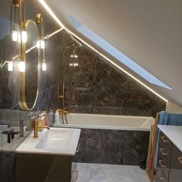 Anna Czapla - Nowoczesna łazienka na poddaszu z czarnymi marmurowymi płytkami, oświetleniem LED i złotymi akcentami, lustro owalne, biała umywalka i wanna.