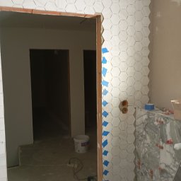 Anna Czapla - Układanie białych, heksagonalnych płytek z subtelnym wzorem na ścianie, zabezpieczonych niebieską taśmą. Obok drewniana ościeżnica, instalacje elektryczne i fragment blatu z marmurowymi płytkami.
