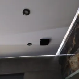 Biały sufit podwieszany z dwoma okrągłymi lampami halogenowymi i czarnym kwadratowym nawiewnikiem wentylacyjnym, widok na fragment ściany z płytek imitujących marmur i szarą ścianę z drzwiami.