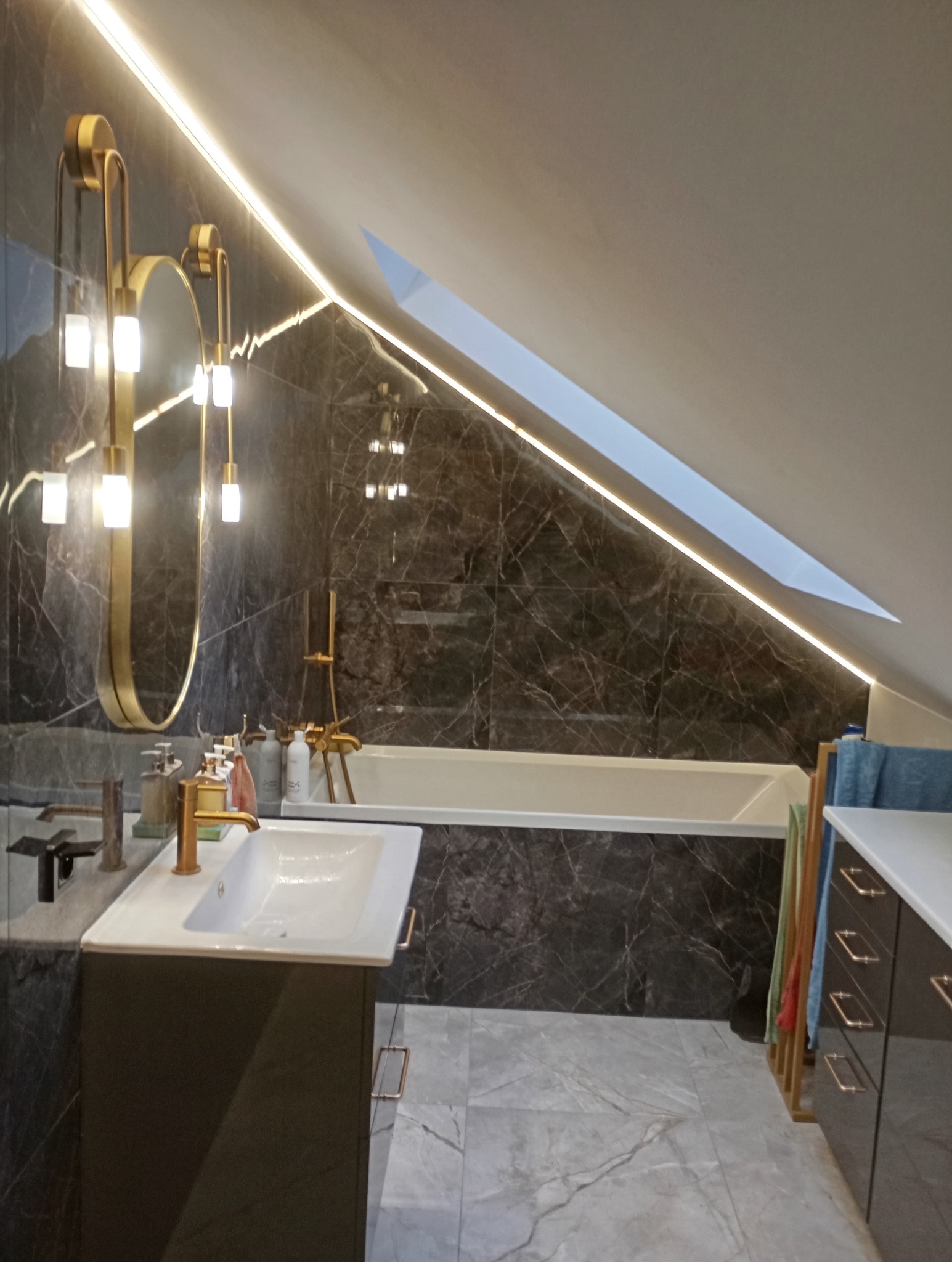 Nowoczesna łazienka na poddaszu z czarnymi marmurowymi płytkami, oświetleniem LED i złotymi akcentami, lustro owalne, biała umywalka i wanna.