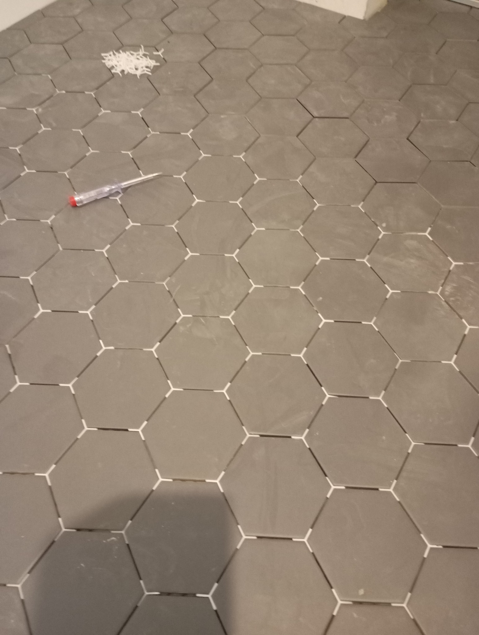 Ułożone szare, heksagonalne płytki podłogowe z białymi krzyżykami dystansowymi, widoczne narzędzie glazurnicze i resztki krzyżyków. Układanie płytek w trakcie.