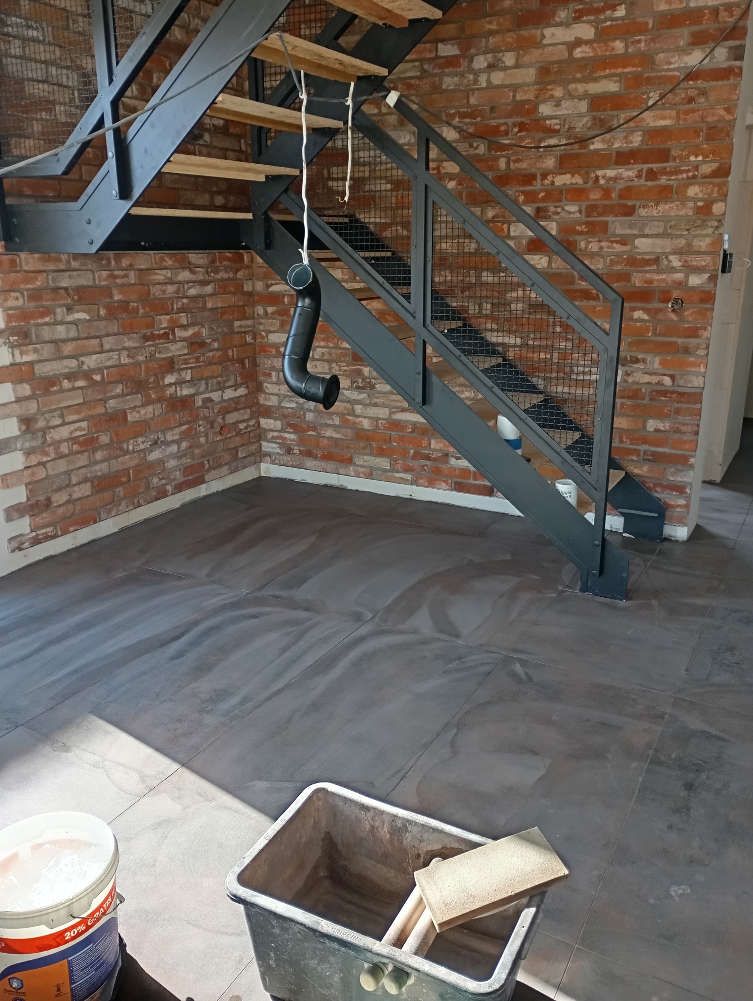 Loftowe wnętrze: ceglana ściana, metalowe schody i szara podłoga z płytek. Wiadro i gąbka sugerują prace wykończeniowe.