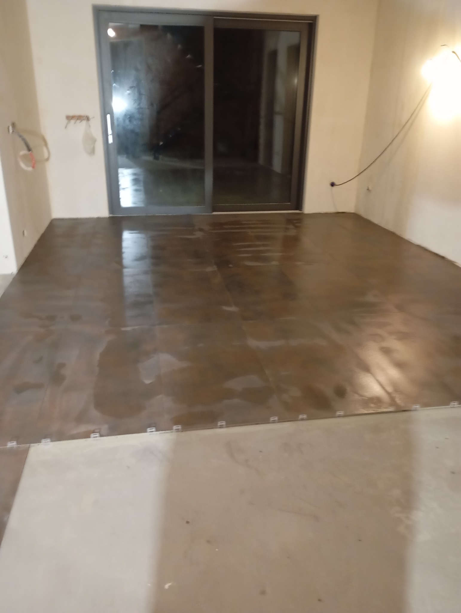 Układanie płytek podłogowych w dużym formacie, z widocznymi krzyżykami dystansowymi. Częściowo ułożona podłoga w surowym wnętrzu z dużym oknem przesuwnym.