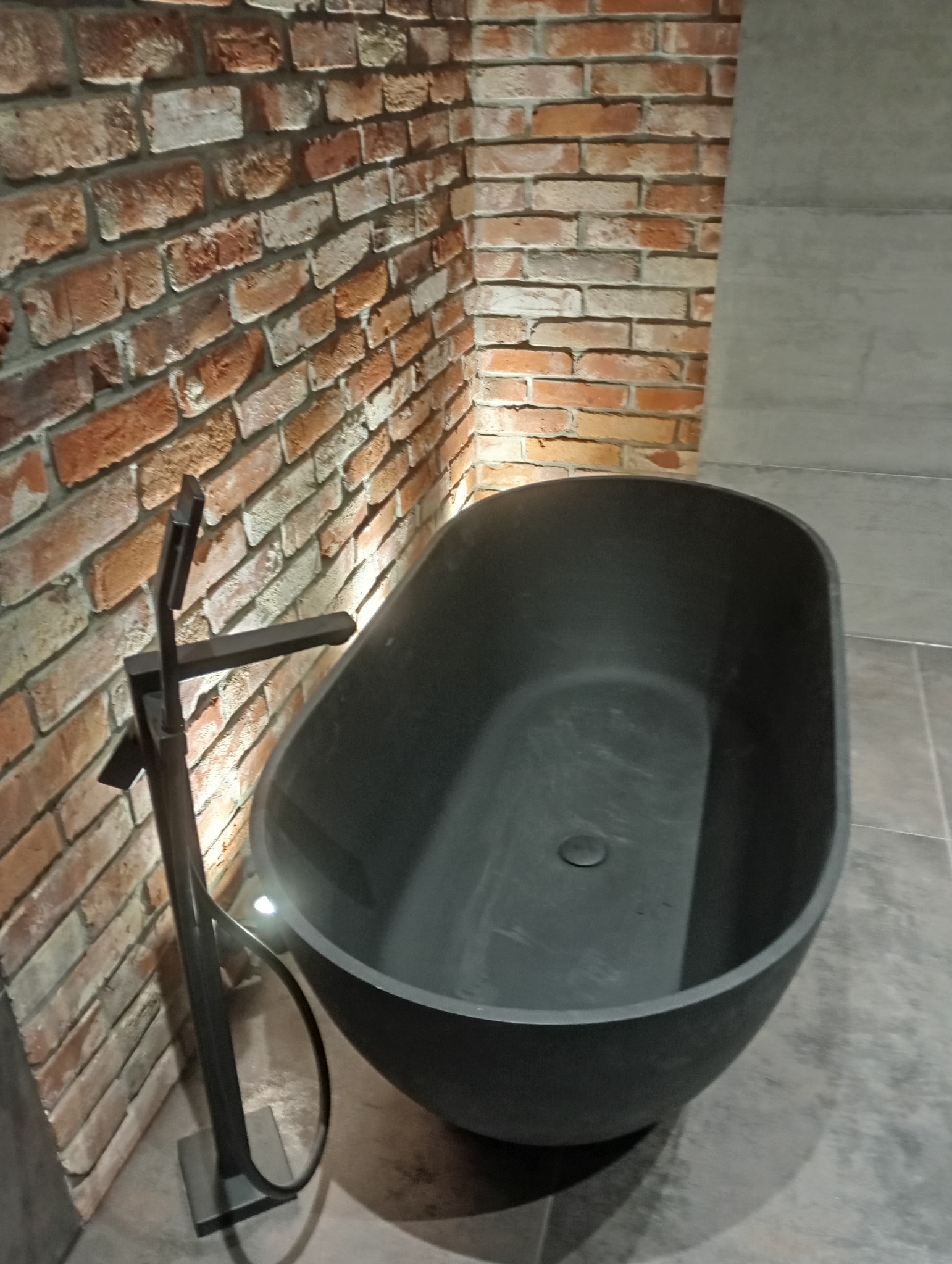 Nowoczesna, wolnostojąca wanna w ciemnym kolorze, z minimalistyczną baterią podłogową. Ściana wykończona czerwoną cegłą i szarą płytą. Stylowe wykończenie łazienki.