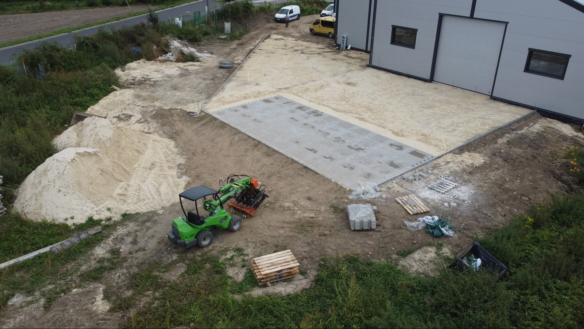 Widok z góry na teren budowy z częściowo ułożoną szarą kostką, zieloną maszyną budowlaną, stertą piasku i paletami. W tle budynek przemysłowy i zaparkowane pojazdy.