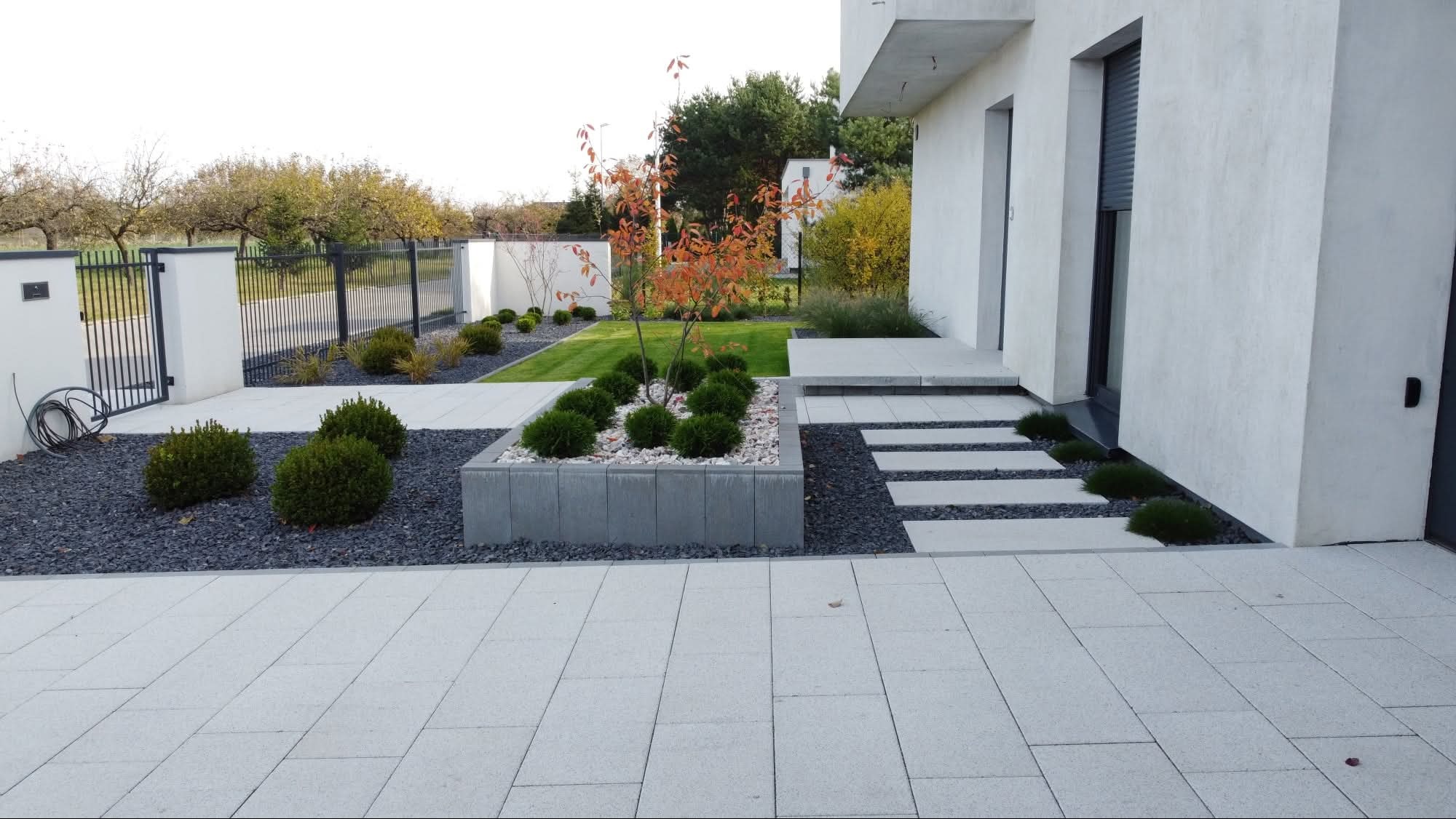 Nowoczesny ogród z jasną kostką, ciemnym żwirem i geometrycznymi rabatami. Minimalistyczny design z elementami zieleni i drzewkiem o pomarańczowych liściach.