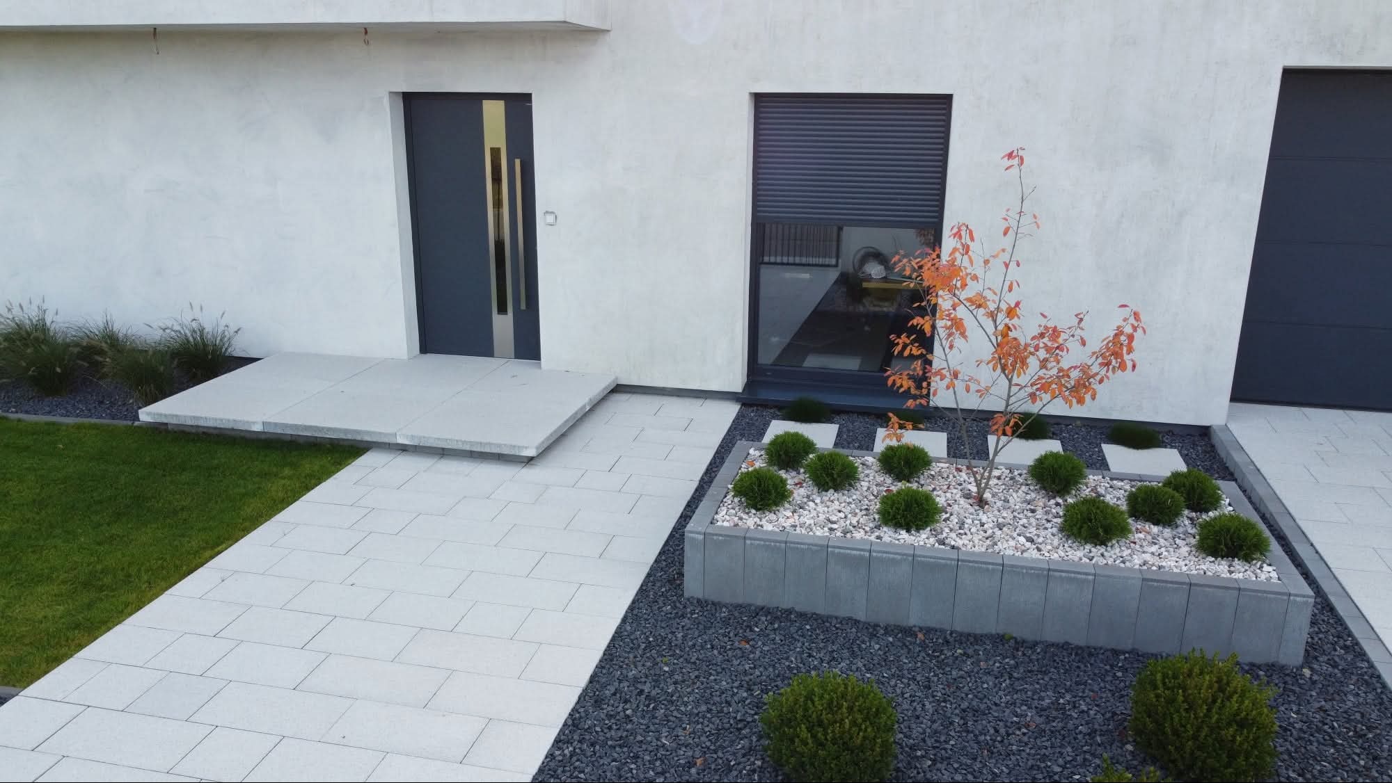 Nowoczesny podjazd z jasnoszarej kostki brukowej prowadzący do domu. Rabata z ozdobnymi kamieniami i krzewami. Minimalistyczny design.