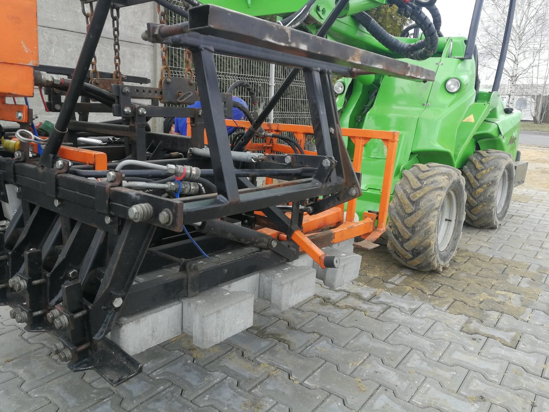 Układanie kostki brukowej maszynowo: zielony traktor z osprzętem do układania kostki, widoczne ułożone fragmenty kostki i paleta z kostką. Praca w toku.