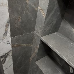 TARABUD - Narożne ujęcie ściany wyłożonej szarymi i białymi płytkami imitującymi marmur, z wnęką wykończoną szarą płytką. Widoczne precyzyjne łączenia płytek.