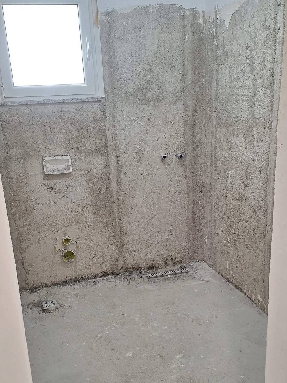 Surowa łazienka w trakcie remontu: ściany z szorstkim tynkiem, widoczne instalacje wodne i odpływ liniowy. Okno z białymi ramami w tle. Etap przygotowania do wykończenia.