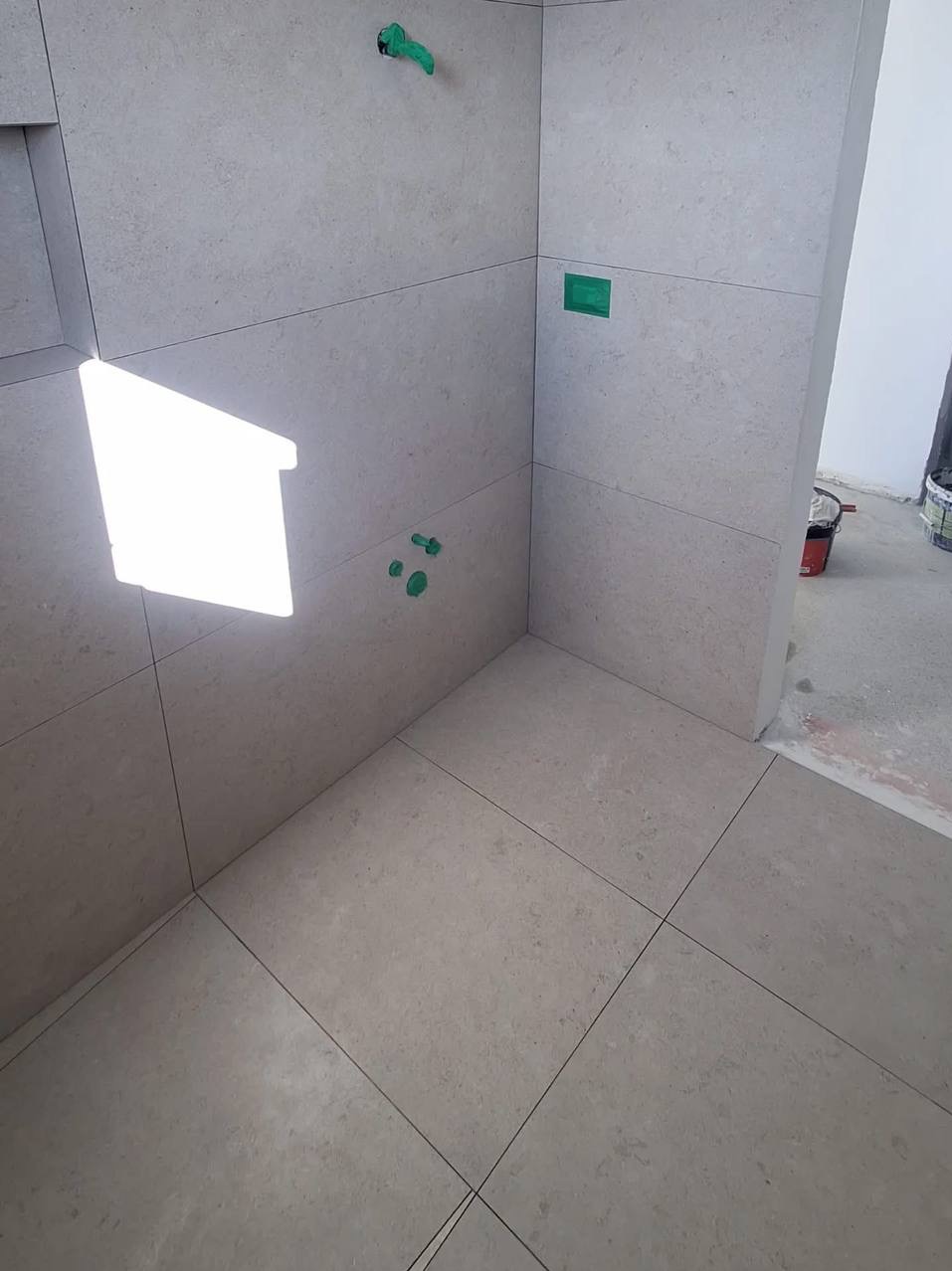 Wykończona łazienka z jasnymi, kwadratowymi płytkami na ścianach i podłodze. Widoczne zielone zaślepki instalacji i fragment pomieszczenia z wiadrami farby.