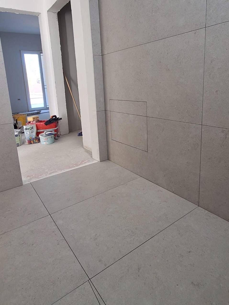 Wykończona ściana i podłoga z dużych, szarych płyt. Widoczny otwór rewizyjny w ścianie. W tle wiaderka z farbami i narzędzia malarskie.