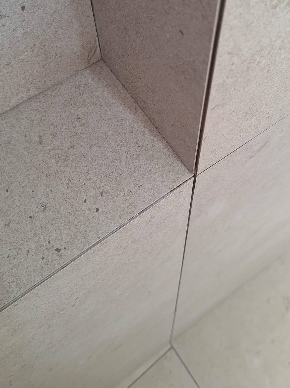 Detaliczne ujęcie narożnika z jasnych, matowych płytek ceramicznych, ukazujące precyzyjne fugi i estetyczne wykończenie wnęki. Minimalistyczny design.