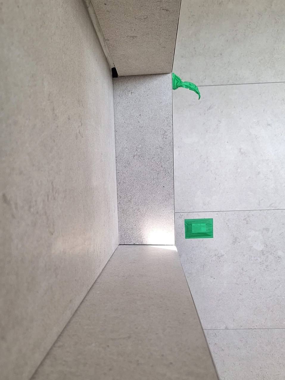 Wykończona ściana i podłoga w łazience z szarych płytek, zabezpieczone zieloną taśmą malarską. Widok z dołu, podkreślający precyzję ułożenia płytek.