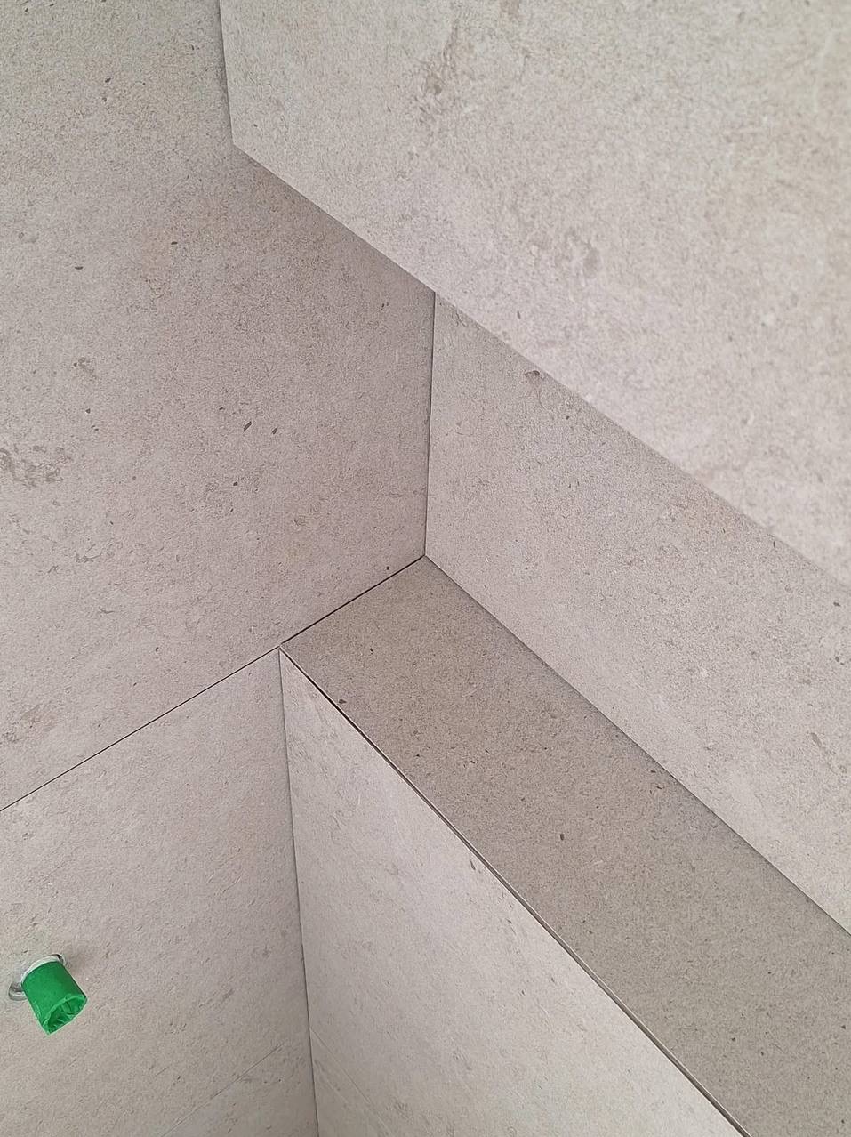 Beżowe, matowe płytki ceramiczne ułożone w narożniku, z widocznym zielonym elementem instalacyjnym w dolnej części kadru. Minimalistyczny design, neutralna kolorystyka.