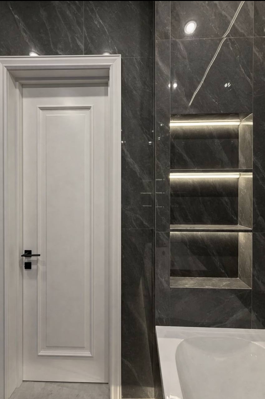 Elegancka łazienka z ciemnymi płytkami imitującymi marmur, podświetlane wnęki i białe drzwi z czarną klamką. Minimalistyczny design i nowoczesne oświetlenie.