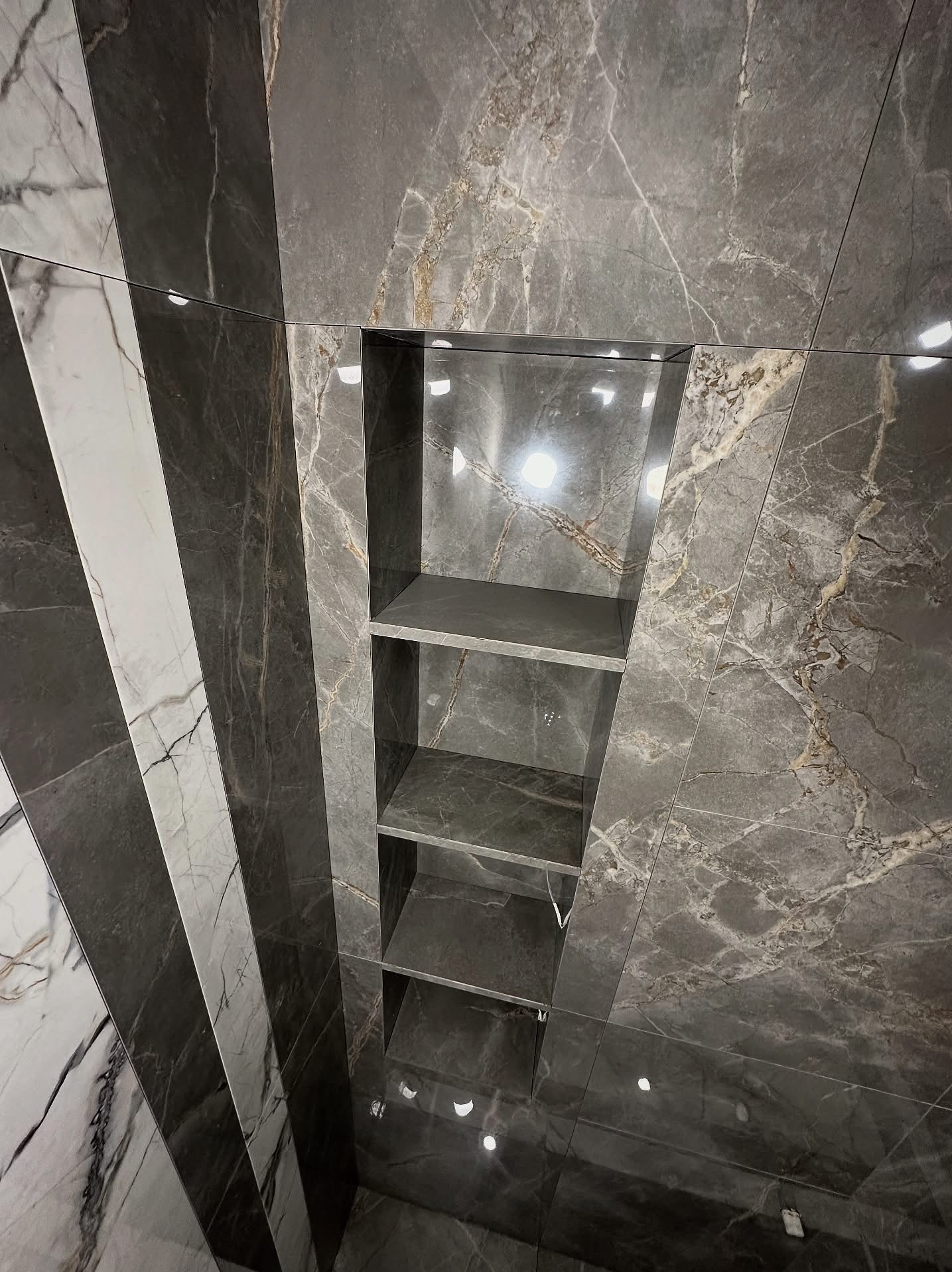 Wykończona ściana łazienki z szarych płytek imitujących marmur, z wnęką z półkami. Ułożenie płytek w pionowe pasy o różnej szerokości i odcieniach szarości.