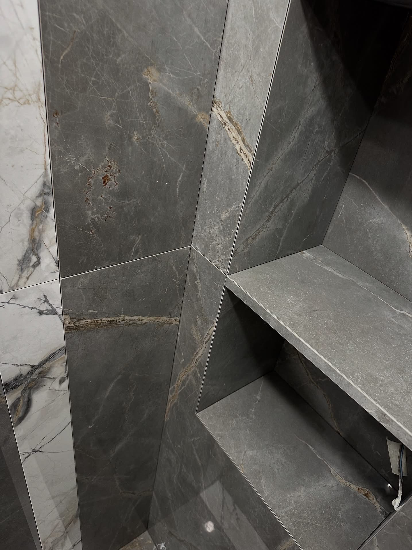 Narożne ujęcie ściany wyłożonej szarymi i białymi płytkami imitującymi marmur, z wnęką wykończoną szarą płytką. Widoczne precyzyjne łączenia płytek.