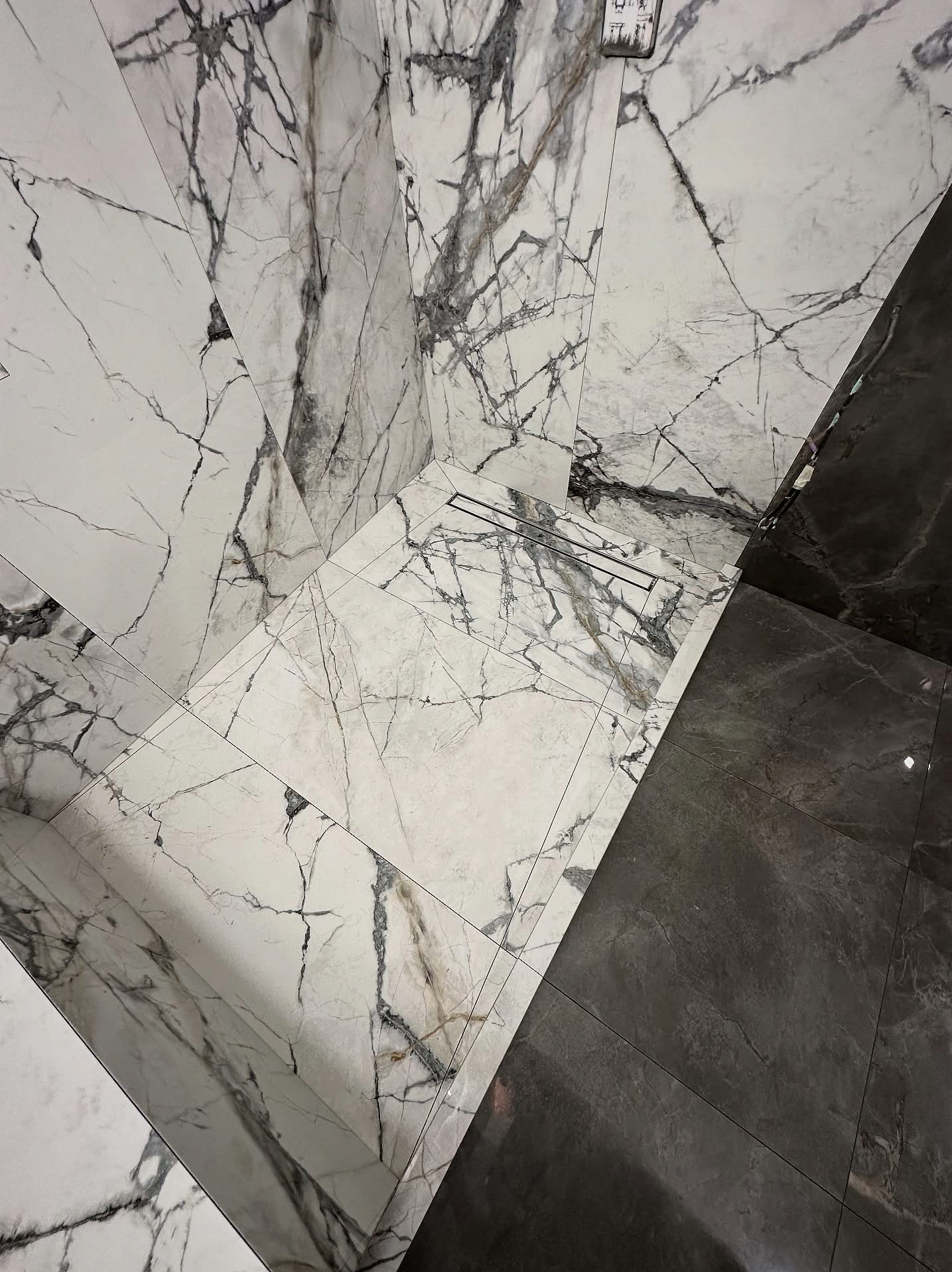 Ukończona podłoga i ściany w łazience wyłożone płytkami imitującymi marmur w odcieniach bieli i szarości. Widoczny odpływ liniowy. Kontrastujące ciemne płytki.