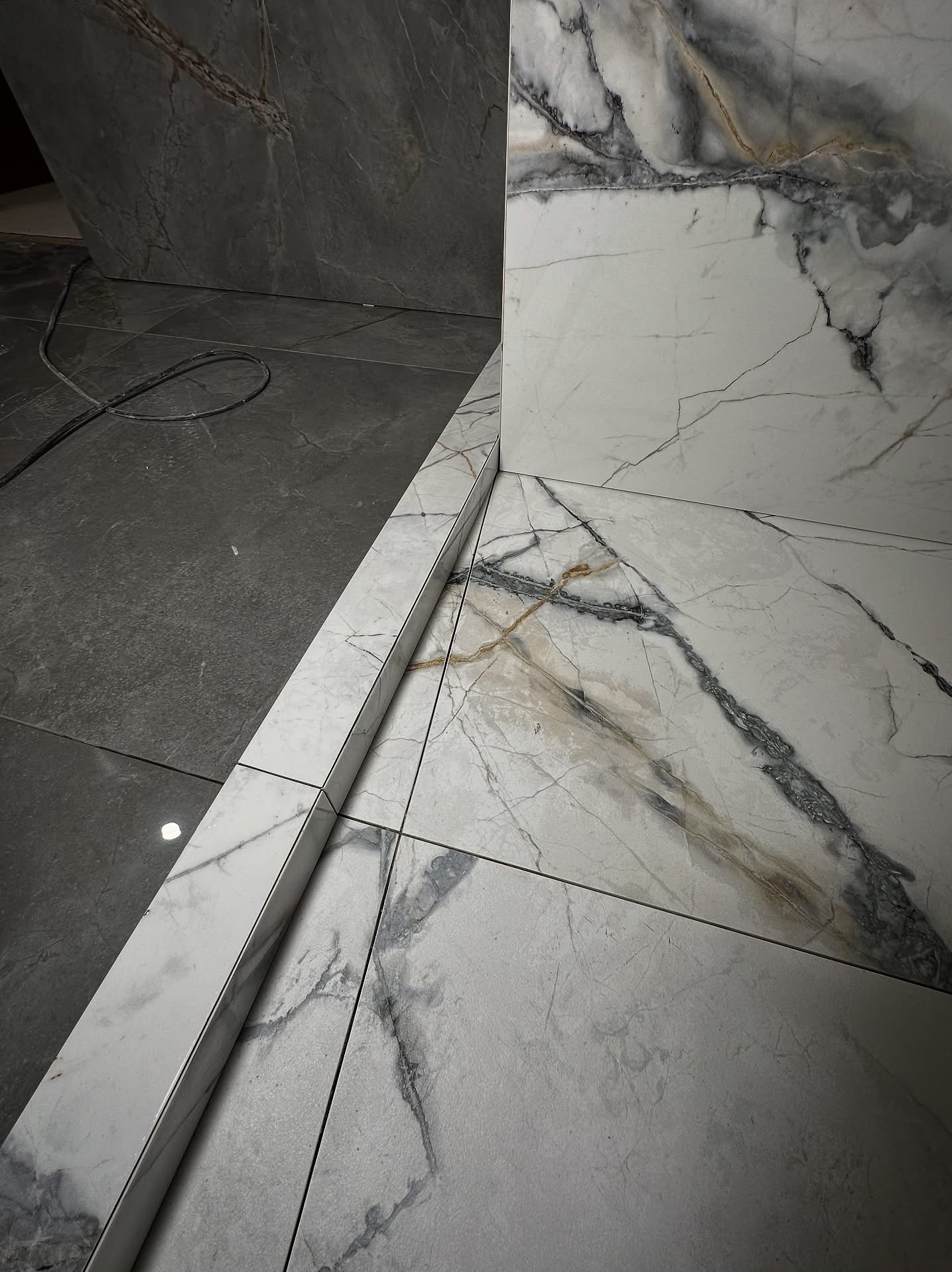 Połączenie dwóch rodzajów płytek: szarej matowej i białej z marmurowym wzorem, oddzielonych listwą. Widok z bliska na podłogę i fragment ściany.