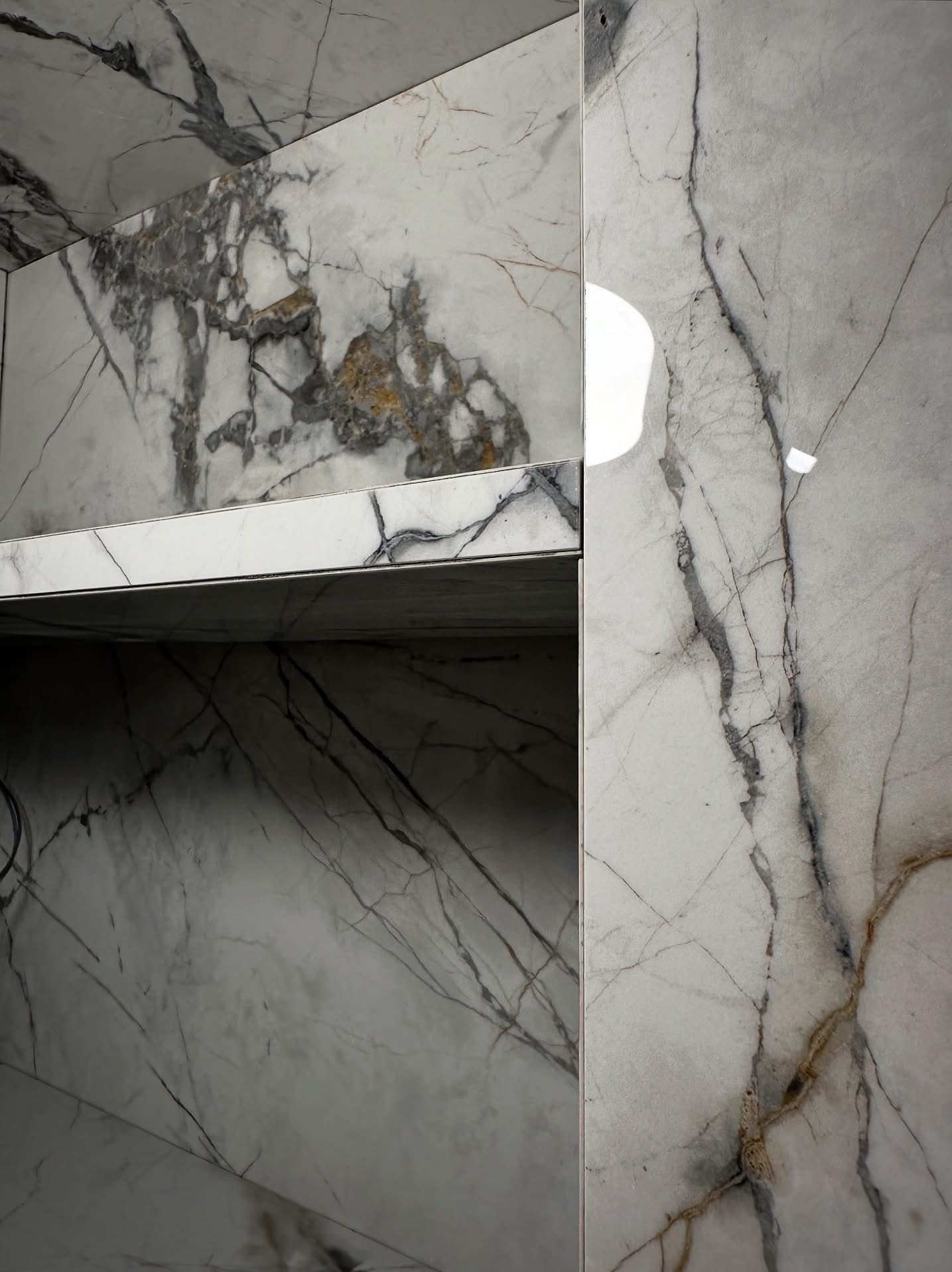 Białe płytki imitujące marmur z szarymi i brązowymi żyłami, ułożone na ścianie i półce w łazience. Widoczne detale wykończenia i połysk powierzchni.