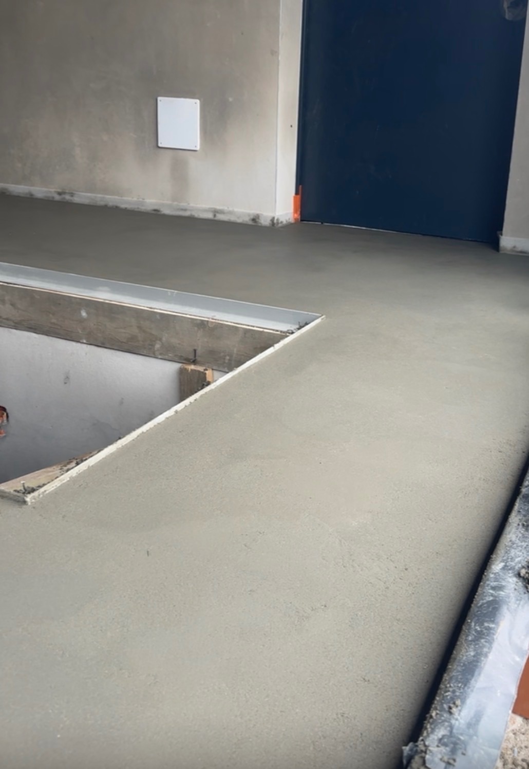 Świeżo wylana, gładka posadzka w szarym odcieniu, z widocznym otworem zabezpieczonym drewnianą ramą. W tle ściana i ciemne drzwi.