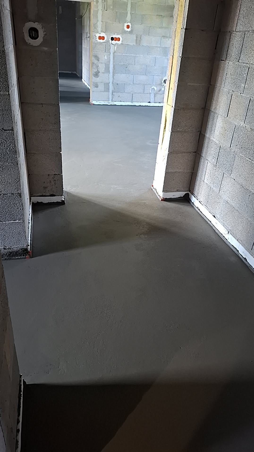 Świeżo wylana, gładka posadzka w surowym wnętrzu budynku z szarego betonu komórkowego. Widoczne listwy dylatacyjne przy ścianach i otwory drzwiowe.