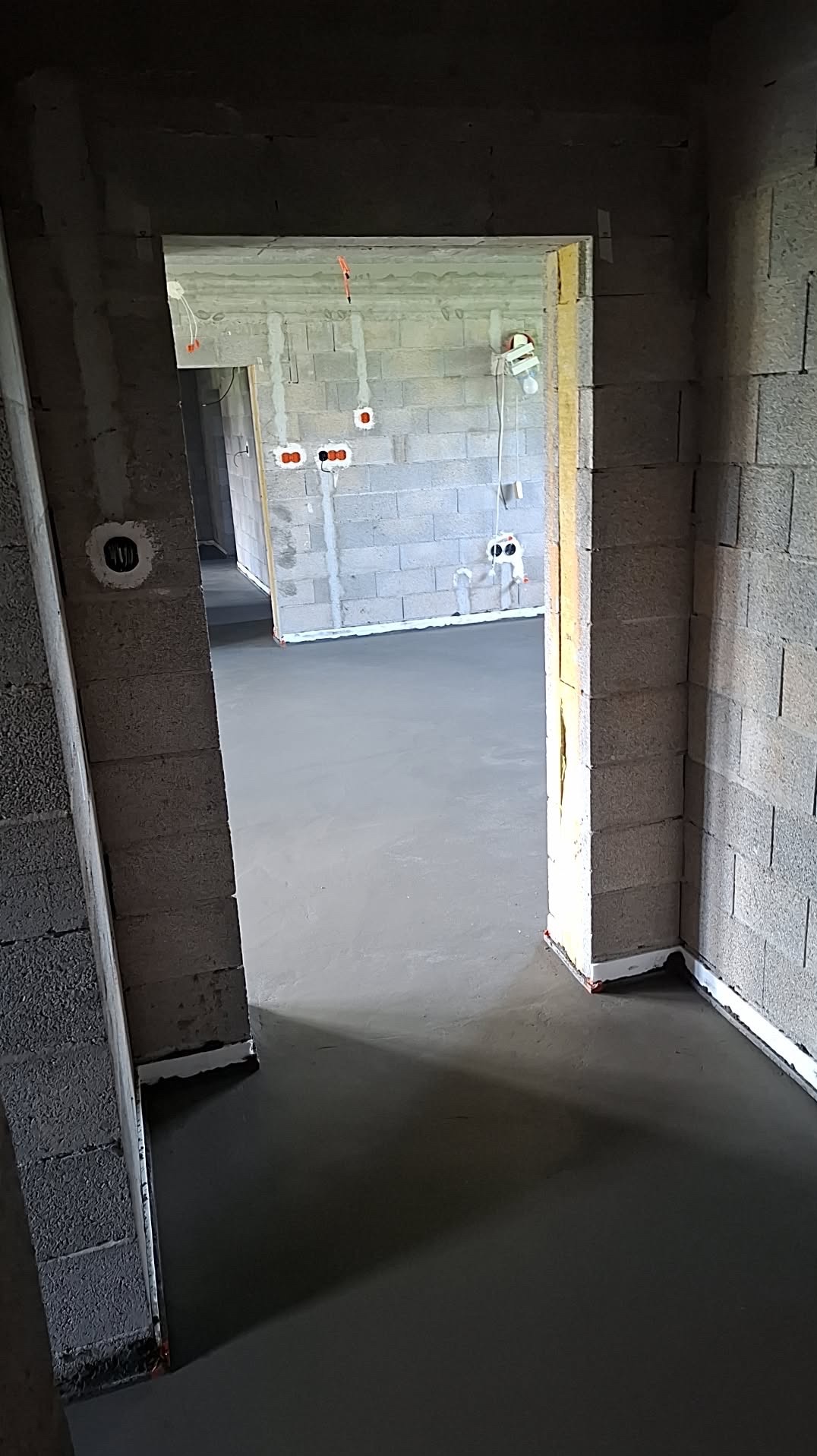 Świeża, gładka wylewka podłogowa w surowym wnętrzu z szarego betonu komórkowego. Widoczne wystające kable elektryczne i biała taśma dylatacyjna przy ścianach.