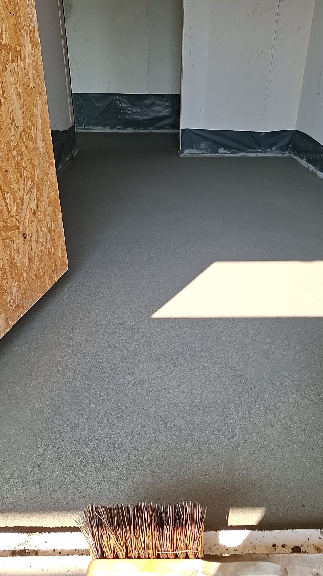 Gładka, szara wylewka w pomieszczeniu z folią przy ścianach. Fragment płyty OSB po lewej i szczotka do zamiatania w dolnej części kadru. Padające światło słoneczne.